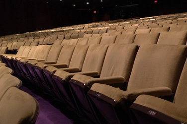 Dimly lighted, several rows of beige chairs in a theater.