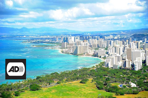 Panoramic view of Honolulu. The turquoise ocean bathes the beaches of the city. A large number of tall buildings rise next to the coast. Superimposed over the image, in the lower left corner, the letters AD appear in black on a white background.