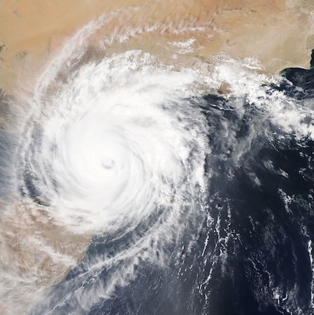 Satelital image. A hurricane spins over a coast.  The hurricane is a large white spiral cloud with long arms. The center of the spiral is dense and as you move away from the center, the arms are less dense.