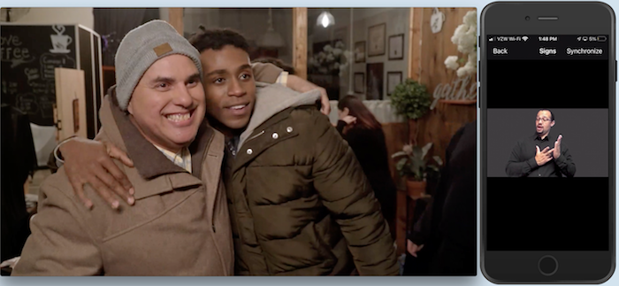 On left, indoors, Robert -a white bald on his 50s with a wool hat- and Steven -a young slim black man- hug each other smiling. Both wear jackets. On right, a phone shows the image of an ASL interpreter making a sign.