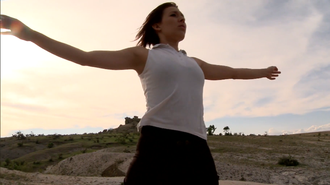 A woman with short hair, wearing a white shirt and a black skirt, stands in a desert with her arms extended at sunrise.