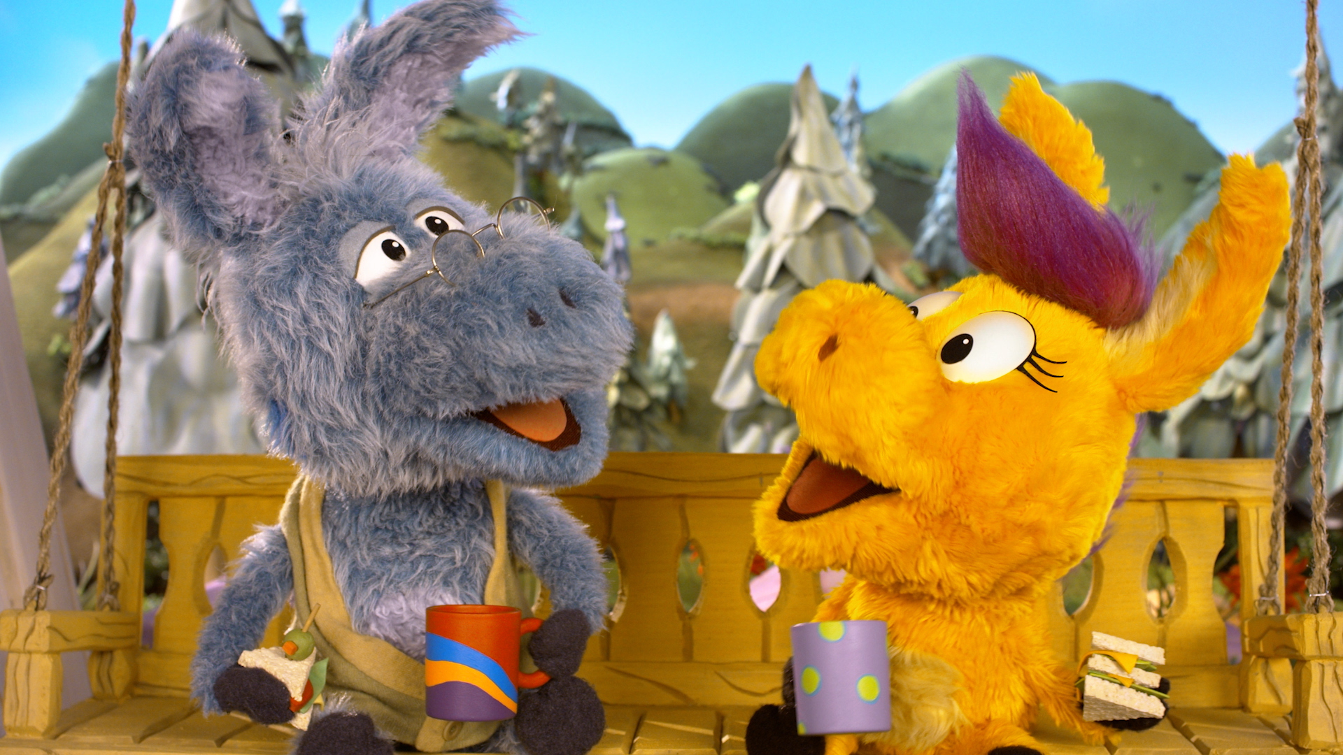 Two Donkey Hodie characters, puppet donkeys, one gray wearing glasses and the other yellow with pink hair, holding cups and sandwiches while chatting. They are seated on a bench against an animated backdrop of trees and mountains.
