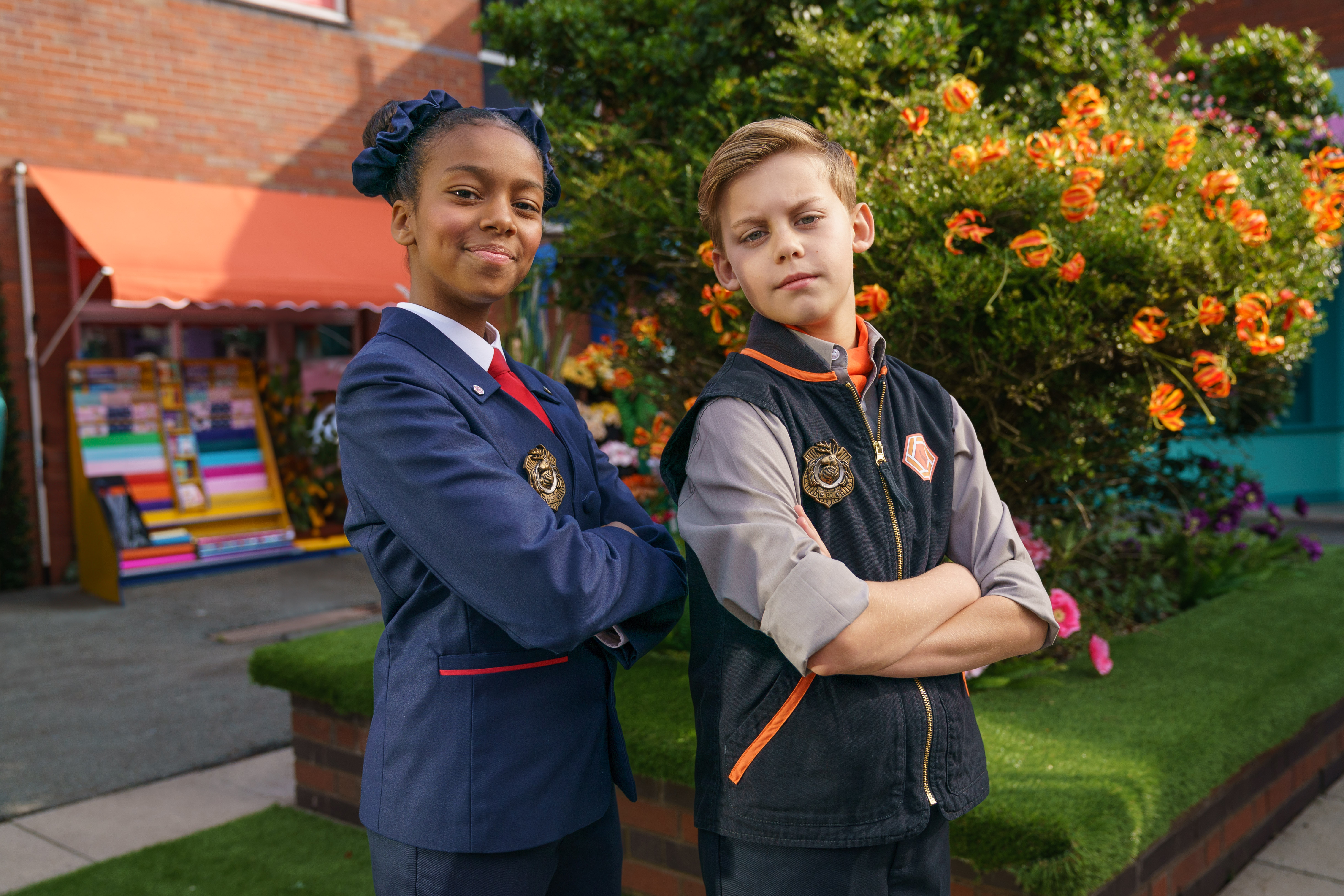 Two teenage children, a boy and a girl, pose in what appears to be a plaza. Both are wearing uniforms with badges and have their arms crossed, looking directly at the camera.