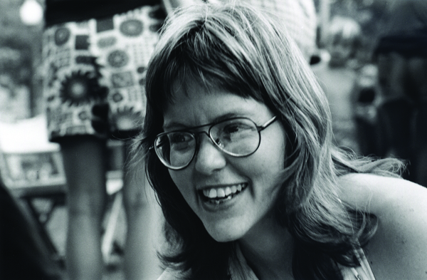 A woman with glasses and a warm smile, captured in a candid black-and-white photograph, with a background suggesting a casual outdoor gathering.