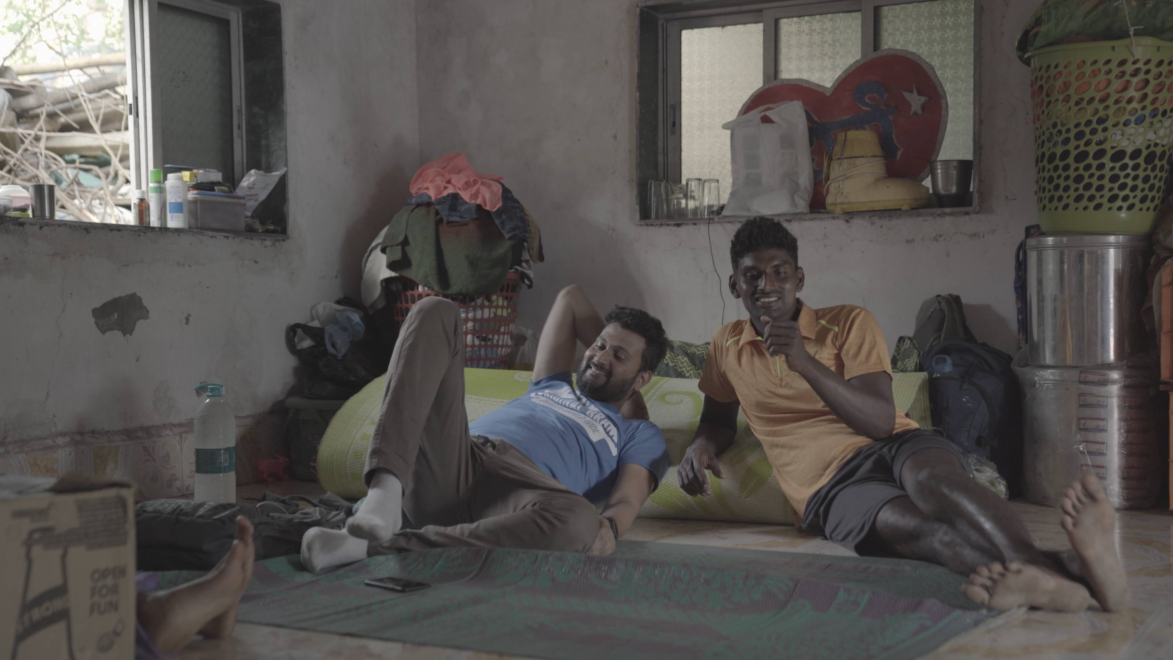 Interiors. Two young Indian men rest on the floor against a pad. They smile and seem to be talking to another person in front of them.