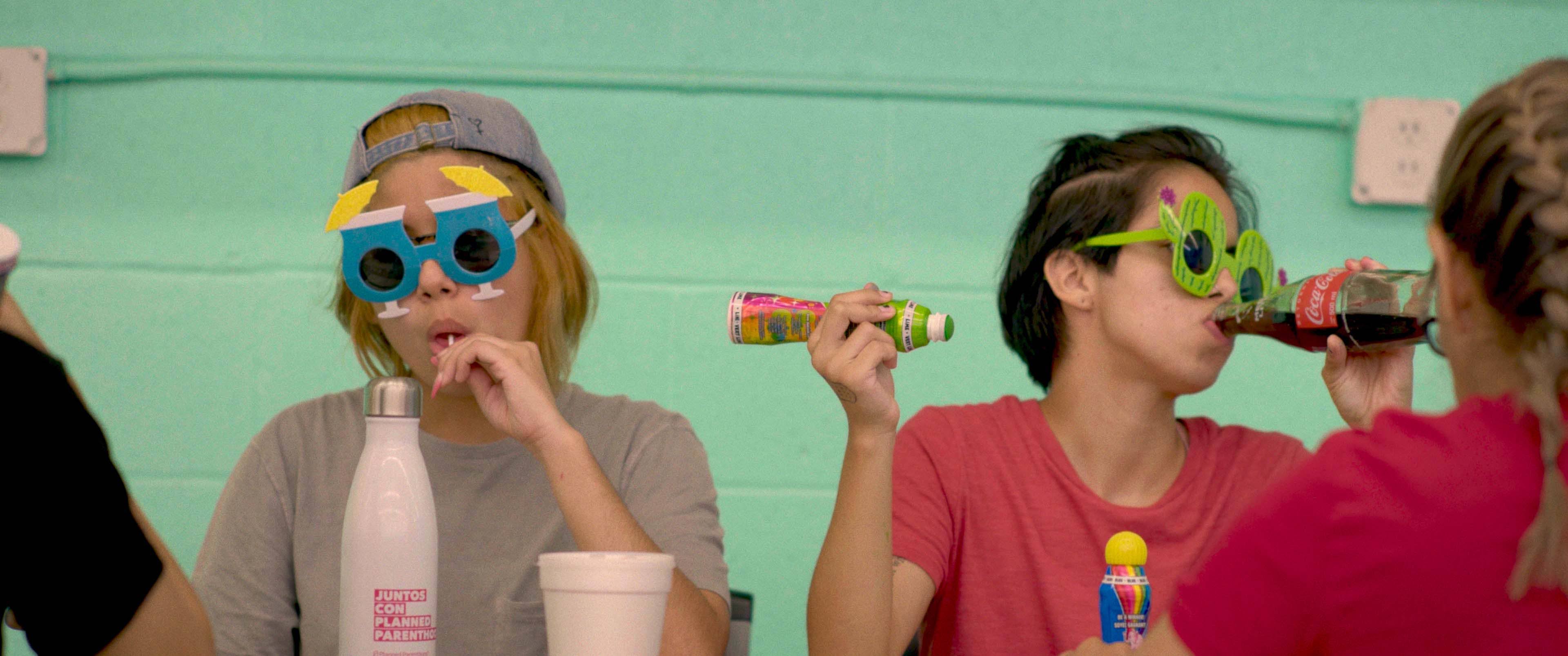 Two girls wearing odd shaped sunglasses are sitting together. The girl on the left enjoys a lollipop, while the girl on the right drinks a soda.