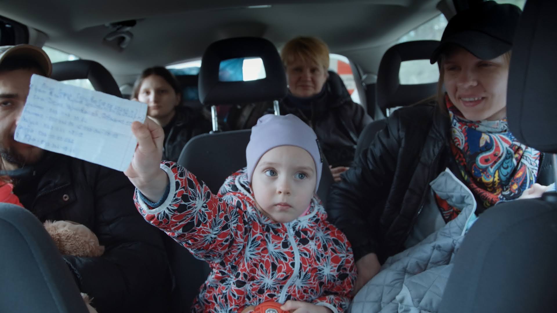 Inside a car, there are four adults and a small girl in the middle holding a piece of paper with a written message. The perspective of the image is as if viewed from the rearview mirror.