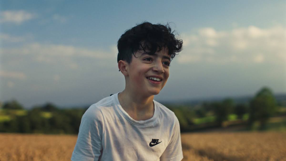 Outdoors, daytime. A smiling boy with curly, dark hair is on an open field under a clear sky.