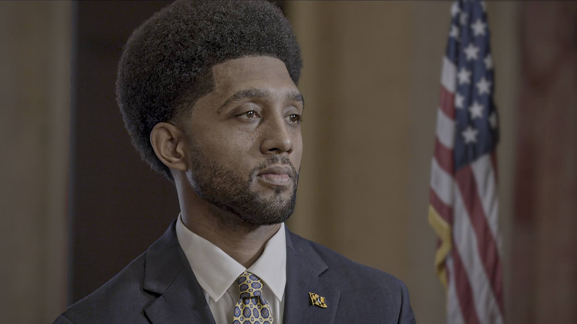 An African American man in formal attire stands looking to the side, with an Afro hairstyle. The blurred United States flag is visible in the background.