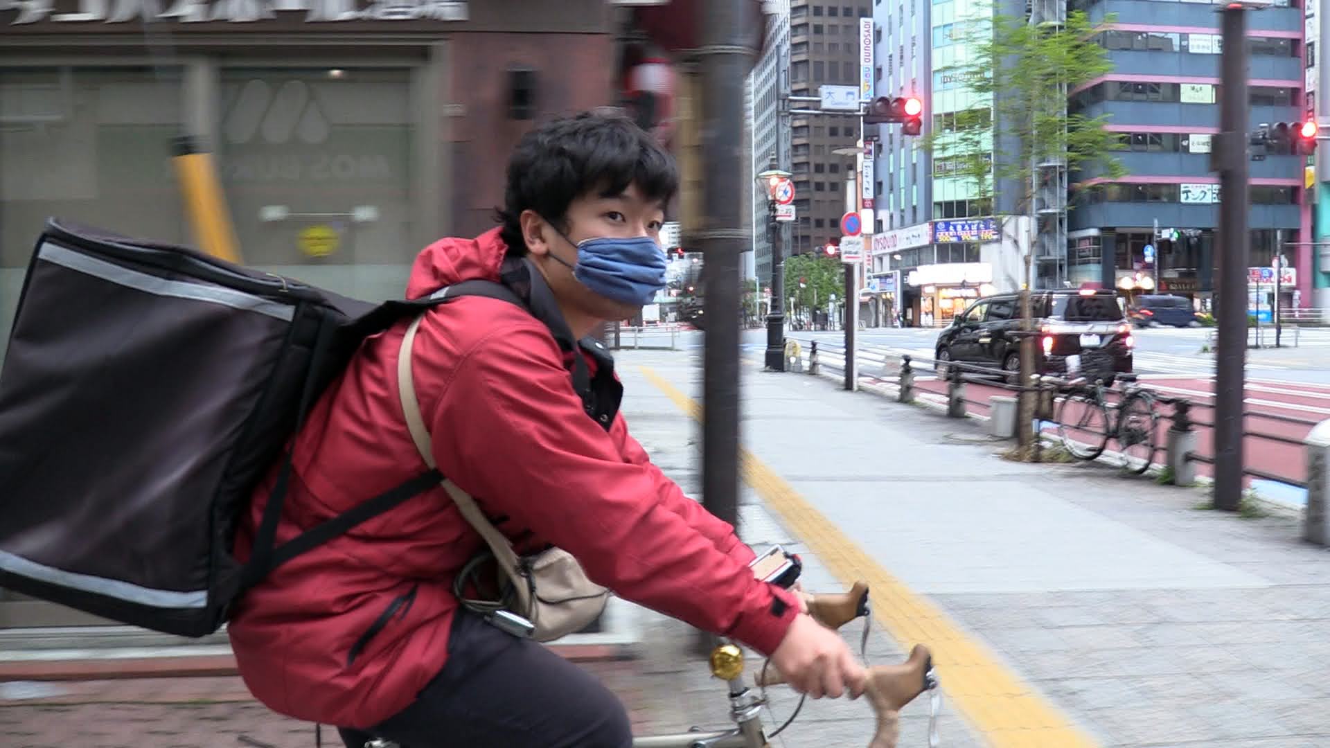 A young man riding a bicycle down a street, wearing a face mask and carrying a delivery backpack.