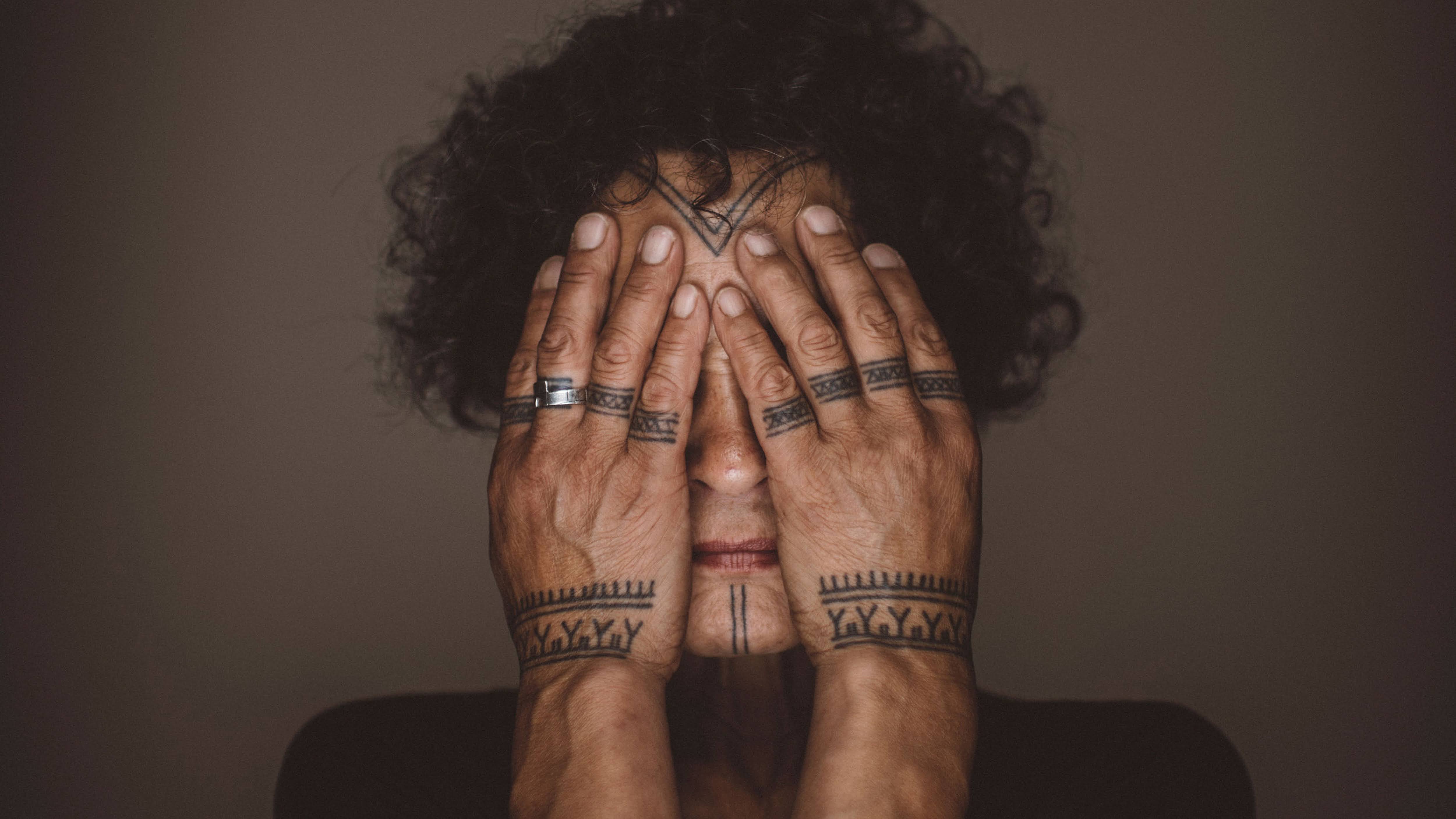 An adult woman with curly black hair covers her face with both hands, revealing several tribal tattoos on her hands and face.