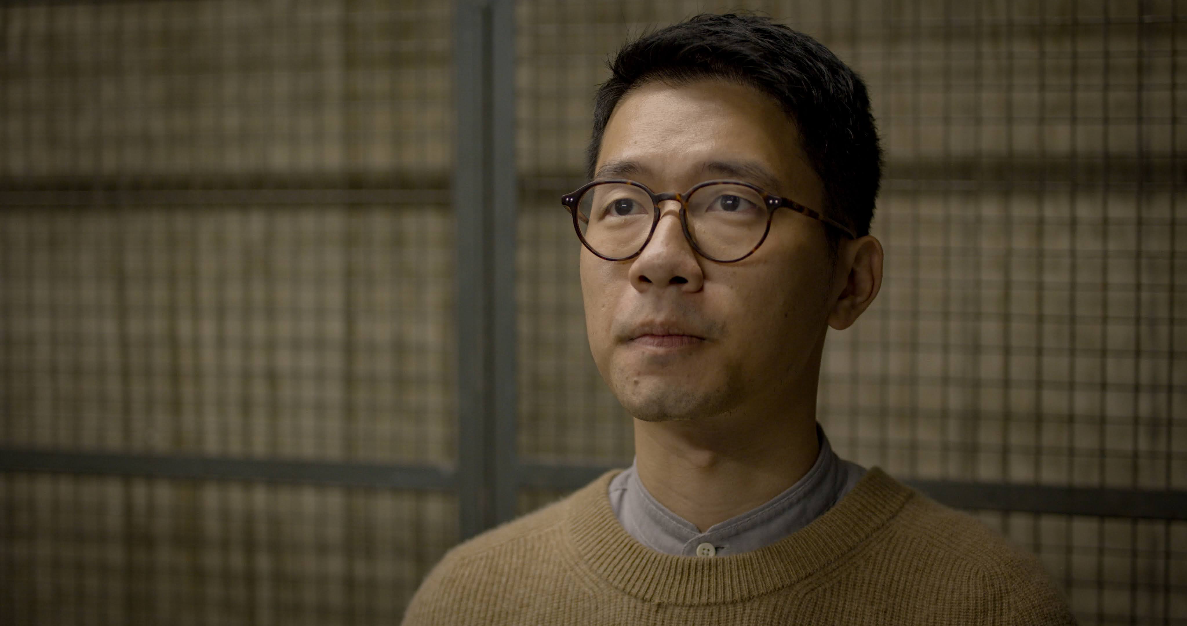 Indoors. A man with round glasses and short hair, with a serious expression, wears a beige sweater over a gray shirt. He looks seriously upwards.