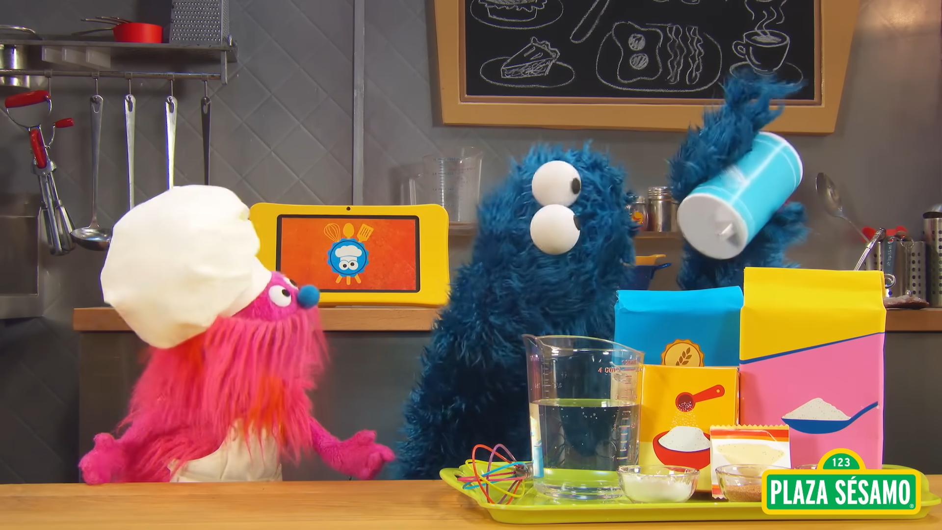 Cookie Monster and Gonger from Sesame Street in a kitchen with ingredients and cooking utensils. Cookie Monster is holding a jar of salt, and Gonger is wearing a chef's hat.