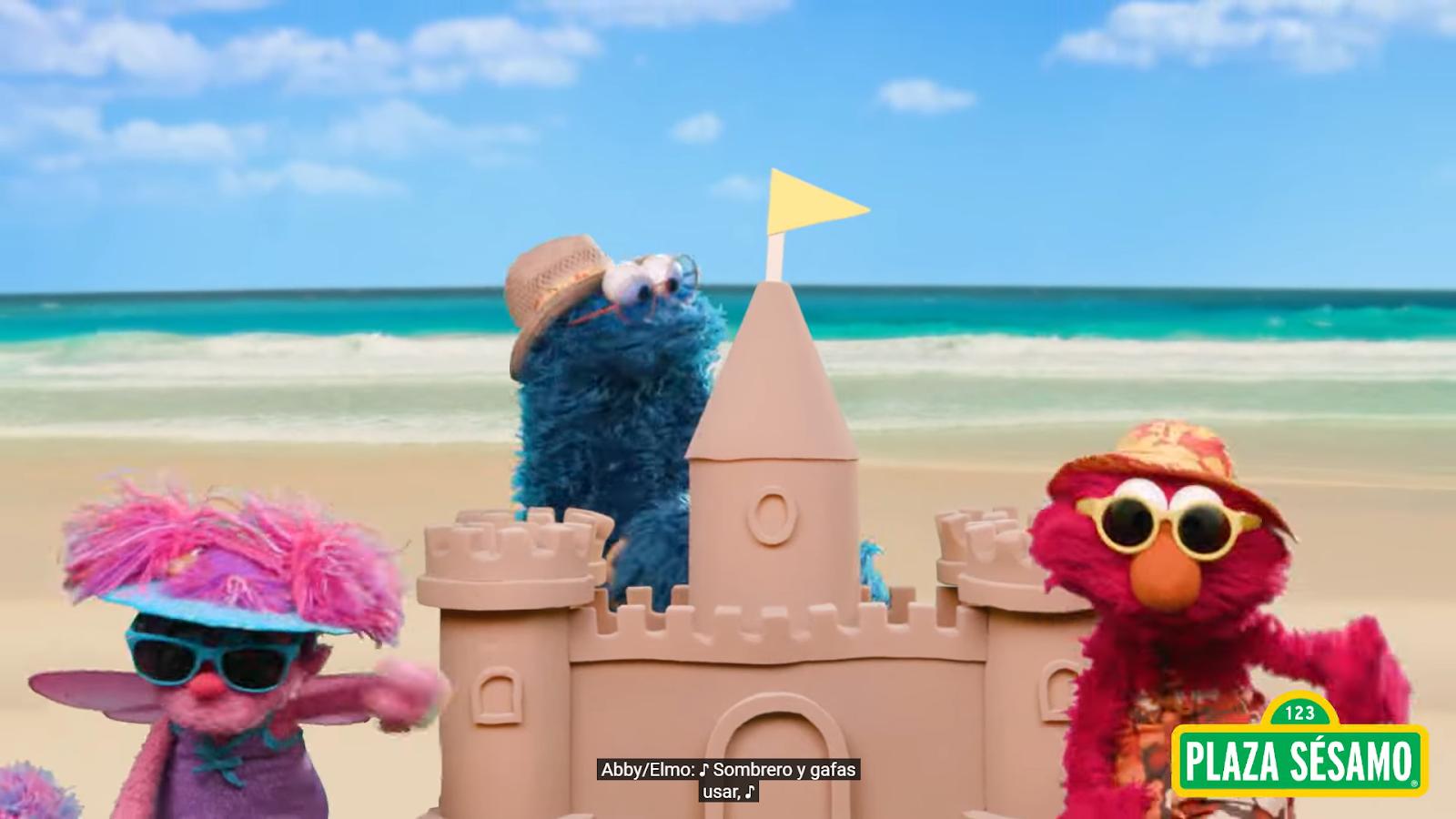 Elmo, Abby, and Cookie Monster at the beach, wearing hats and sunglasses, with a sandcastle between them.