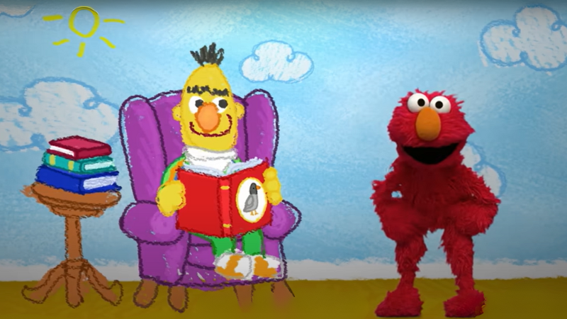 Elmo stands next to Beto, who is seated in a chair reading books