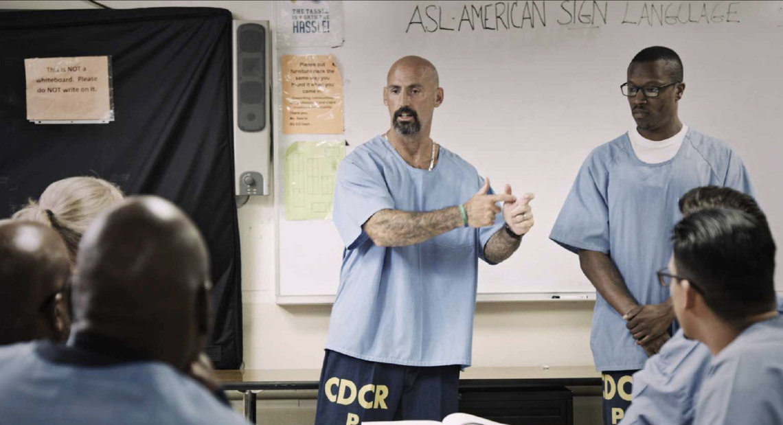 A bald man with a beard stands in a room with seated people, gesturing with his hands. Next to him, another man is standing, and in the background on a board, the phrase ASL American Sign Language is visible.