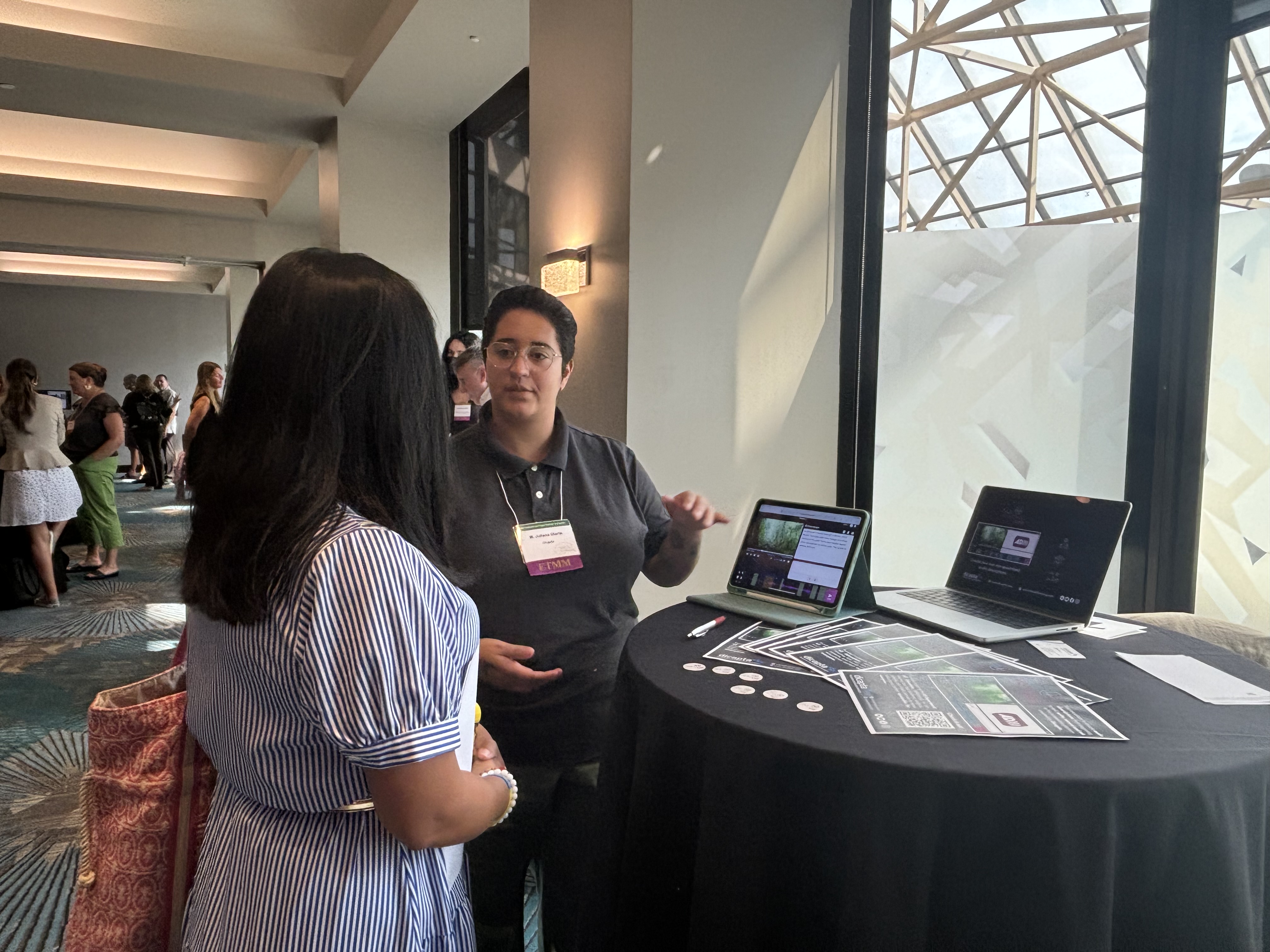 Indoors. Juliana Olarte of Dicapta's team talking to a woman about All4Voicing Lite beside a display table at a conference, featuring two laptops showcasing All4Voicing Lite, along with flyers and buttons.