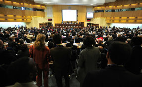 Conference room. A large audience watches a group of panelists.